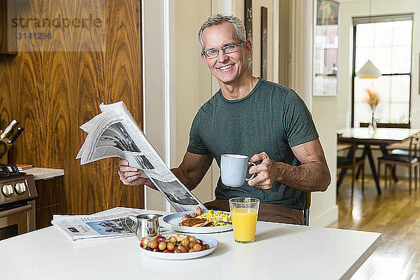 Porträt eines lächelnden  reifen Mannes  der am Esstisch sitzt und Kaffeetasse und Zeitung hält