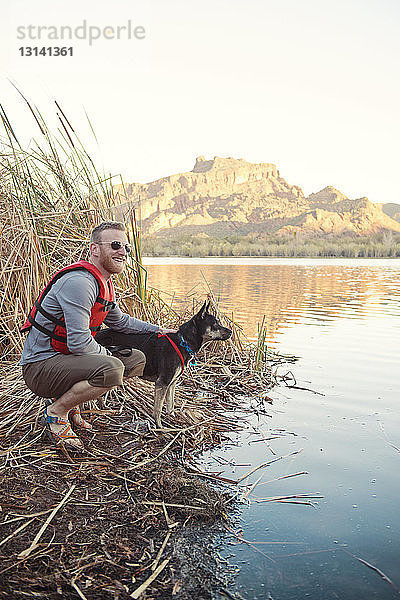 Seitenansicht eines lächelnden Mannes  der mit Hund am Seeufer kauert