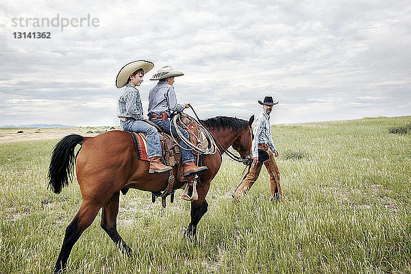 Vater schaut auf Söhne  die auf einem Pferd auf einem Feld vor bewölktem Himmel reiten