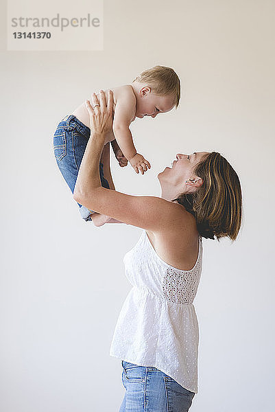 Seitenansicht einer spielerischen Mutter  die ihren hemdlosen Sohn vor weißem Hintergrund anhebt