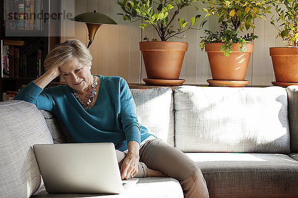 Frau mit der Hand im Haar  die einen Laptop benutzt  während sie zu Hause auf dem Sofa sitzt