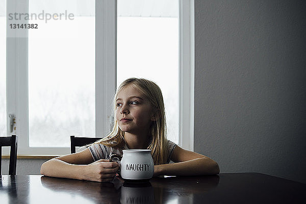 Nachdenkliches Mädchen hält Becher  während es zu Hause auf einem Stuhl am Tisch sitzt