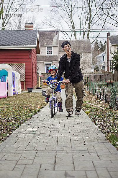 Glücklicher Vater hilft Sohn beim Fahrradfahren auf Fußweg im Hinterhof