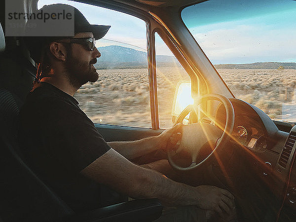 Seitenansicht eines selbstbewussten Mannes mit Sonnenbrille und Mütze beim Fahren eines Wohnmobils bei Sonnenuntergang