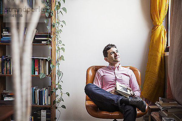 Nachdenklicher Mann hält Buch  während er zu Hause auf einem Stuhl sitzt