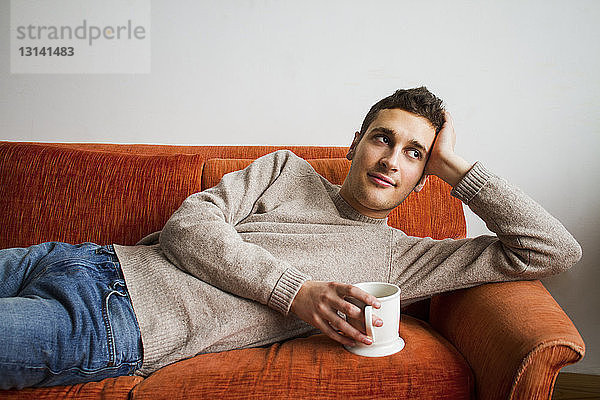 Nachdenklicher Mann hält Kaffeetasse  während er zu Hause auf dem Sofa liegt
