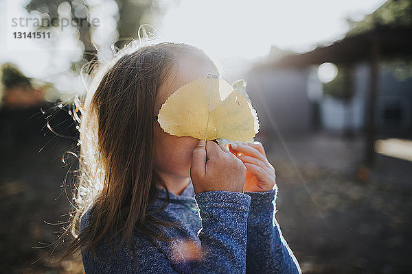 Süßes Mädchen versteckt Gesicht mit Blättern an einem sonnigen Tag