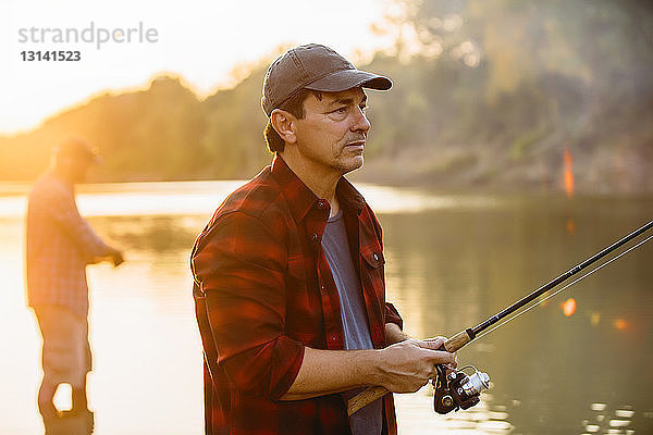 Mittelgroßer erwachsener Mann schaut weg  während sein Freund bei Sonnenuntergang im Hintergrund fischt