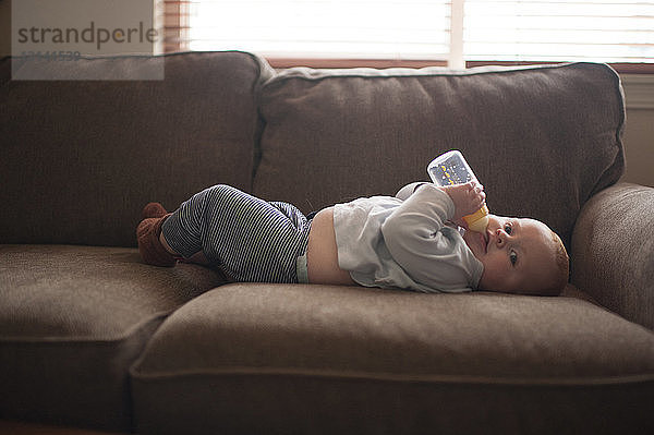 Porträt eines kleinen Jungen  der Milch aus der Flasche trinkt  während er zu Hause auf dem Sofa liegt