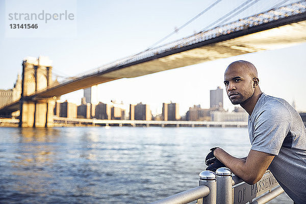 Seitenansicht eines nachdenklichen männlichen Athleten  der auf der Promenade mit der Brooklyn Bridge im Hintergrund steht
