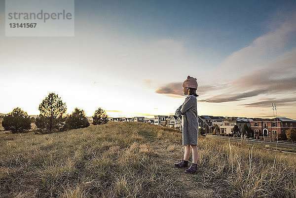 Seitenansicht eines Mädchens  das bei Sonnenuntergang auf einem Grasfeld gegen Himmel und Stadtbild steht
