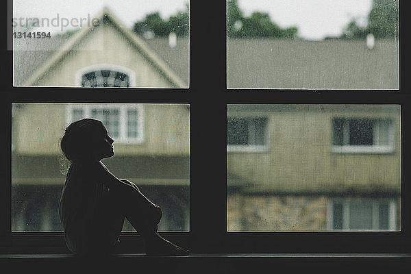 Nachdenkliches Mädchen sitzt auf Fensterbank gegen Haus