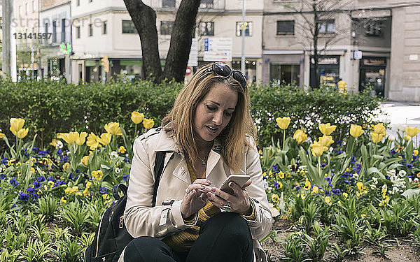 Geschäftsfrau benutzt Mobiltelefon  während sie an Pflanzen in der Stadt sitzt