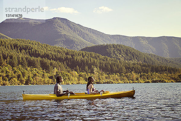 Seitenansicht eines im Kanu sitzenden Paares auf einem Fluss gegen einen Berg