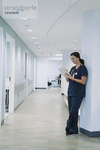Ärztin in voller Länge am Tablet-Computer  während sie sich im Krankenhaus an eine architektonische Säule lehnt