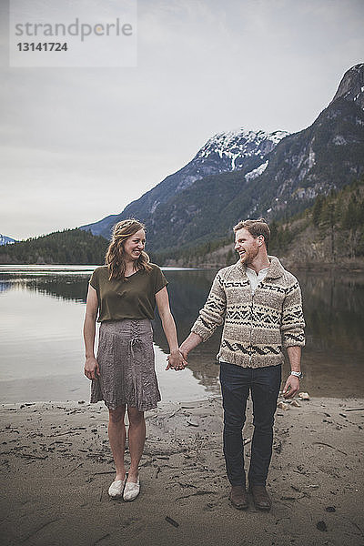 Lächelndes junges Paar in voller Länge hält sich an den Händen  während es am Seeufer im Silver Lake Provincial Park steht