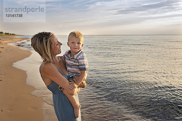 Porträt eines süßen Sohnes  der von der Mutter bei Sonnenuntergang am Strand getragen wird