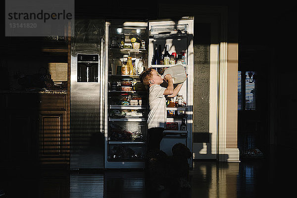 Seitenansicht eines Jungen  der Milch aus einer Flasche trinkt  während er zu Hause in der Dunkelkammer gegen einen offenen Kühlschrank steht