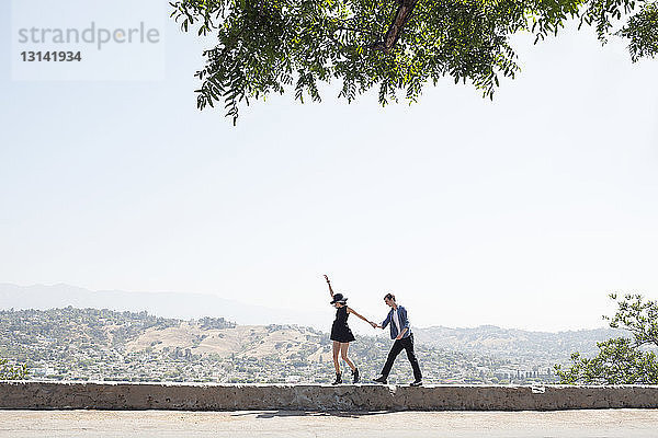 Aufgeregtes junges Paar geht auf Stützmauer gegen klaren Himmel