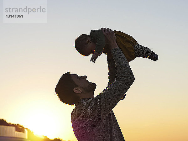 Tiefwinkelansicht eines glücklichen Vaters  der seine Tochter aufhebt  während er bei Sonnenuntergang am klaren Himmel steht