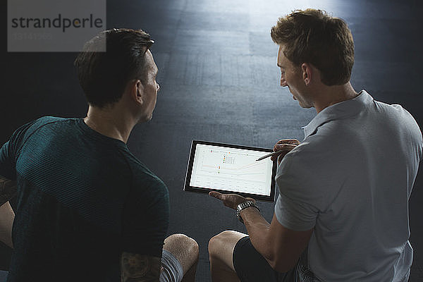 Hochwinkelansicht des Trainers  der dem Kunden einen Tablet-PC zeigt  während er im Fitnessstudio sitzt