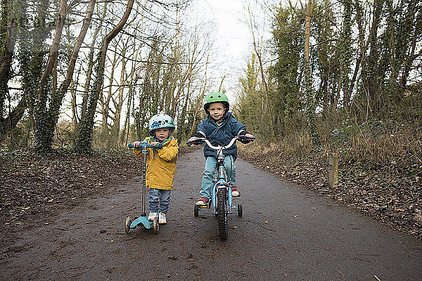 Portrait von Geschwistern beim Schieben von Roller und Fahrrad auf der Strasse gegen den Himmel
