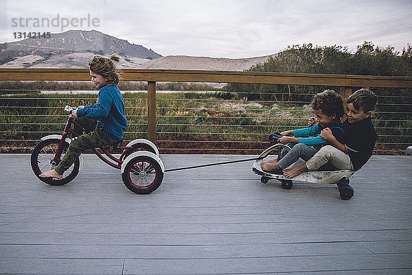 Junge zieht Spielzeugauto mit Freunden  während er auf dem Bodenbrett Dreirad fährt