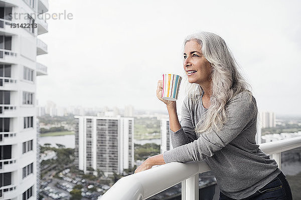 Nachdenkliche reife Frau trinkt Kaffee  während sie auf dem Balkon steht