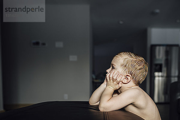 Seitenansicht eines nachdenklichen kleinen Jungen mit Händen am Kinn  der zu Hause am Tisch sitzt