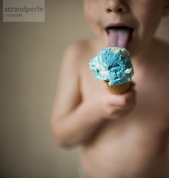 Mittelteil eines Jungen ohne Hemd  der Eiscreme leckt  während er zu Hause an der Wand steht