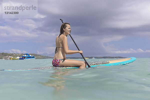 Frau im Bikini paddelt auf dem Meer vor bewölktem Himmel