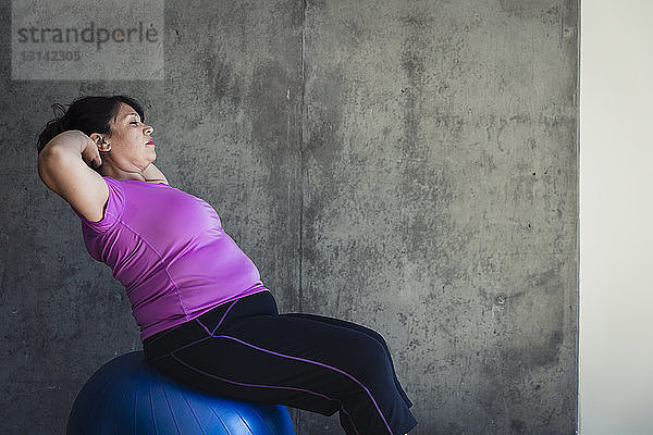 Frau mit Händen hinter dem Kopf sitzend auf Fitnessball gegen Wand im Yogastudio