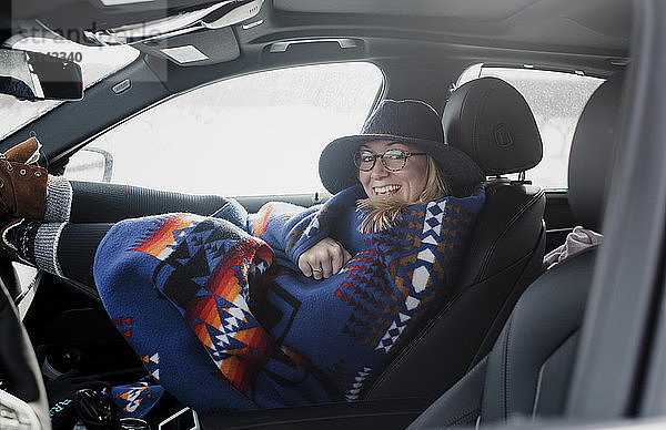 Porträt einer glücklichen  in eine Decke gehüllten Frau  die im Auto sitzend durch das Fenster schaut