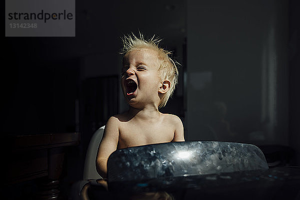 Kleiner Junge ohne Hemd schreit  während er die Brise der Klimaanlage in der Dunkelkammer zu Hause genießt