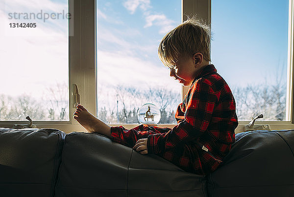 Seitenansicht eines Jungen  der auf Dekor schaut  während er zu Hause auf einem Sofa am Fenster sitzt