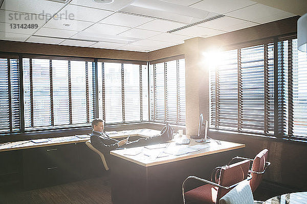 Geschäftsmann benutzt Mobiltelefon  während er sich an einem sonnigen Tag im Büro entspannt