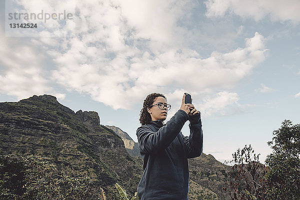 Niedrigwinkelansicht einer Frau  die mit einem Mobiltelefon fotografiert  während sie am Berg steht