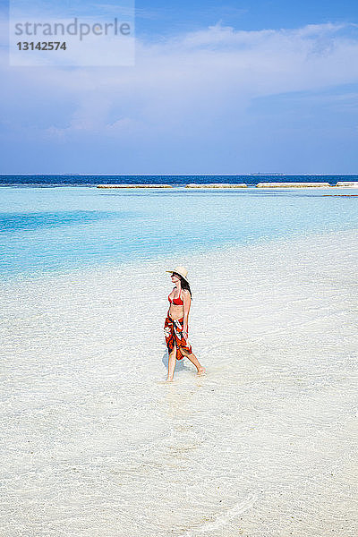 Schrägansicht einer Frau mit Bikini und Hut beim Spaziergang am Strand gegen den Himmel während eines sonnigen Tages