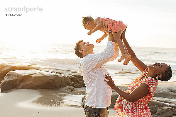 Glückliche Eltern spielen mit ihrer Tochter  während sie am Strand gegen den Himmel stehen