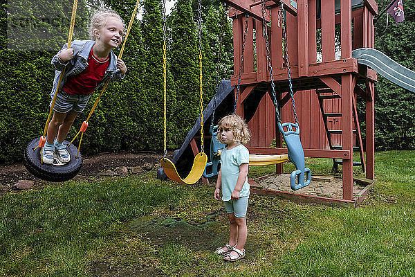 Trauriges Mädchen schaut die glückliche Schwester an  die auf einer Reifenschaukel auf dem Spielplatz schaukelt