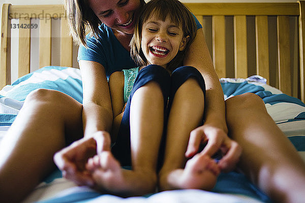 Glückliche Mutter kitzelt Tochter beim Sitzen im Bett an den Füßen