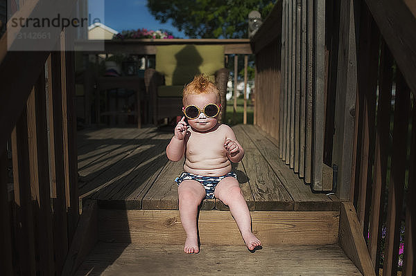 Porträt eines Jungen ohne Hemd mit Sonnenbrille auf der Veranda sitzend