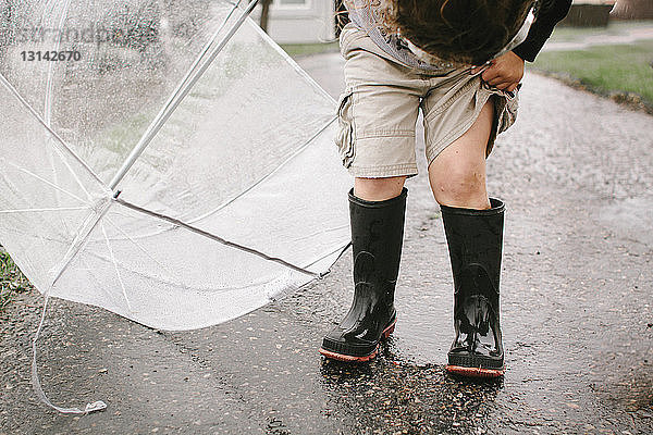 Mädchen in Gummistiefeln hält Regenschirm  während sie auf nasser Straße steht