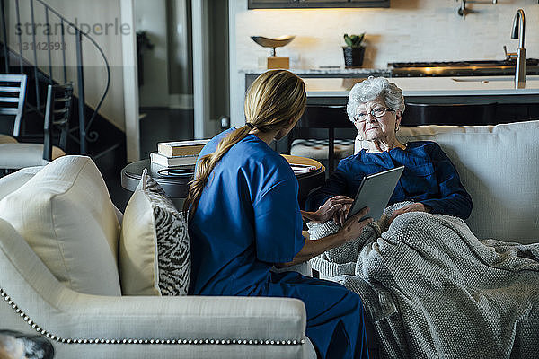Hausangestellte diskutiert über Tablet-Computer mit älterer Frau im Wohnzimmer