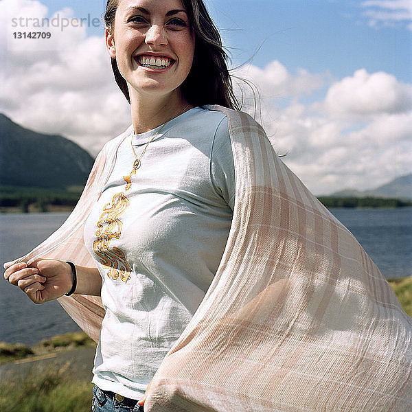 Fröhliche Frau hält Schal  während sie am Seeufer vor bewölktem Himmel steht