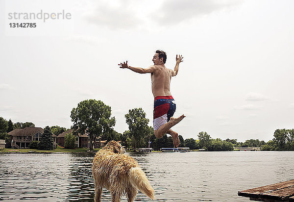 Aufgeregter Mann in voller Länge mit Hund springt im See gegen den Himmel