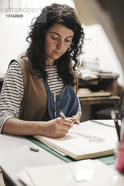 Weibliche Kunsthandwerkerin zeichnet auf Buch  während sie im Workshop sitzt