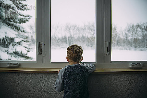 Rückansicht eines Jungen  der durch ein Fenster schaut  während er im Winter zu Hause steht