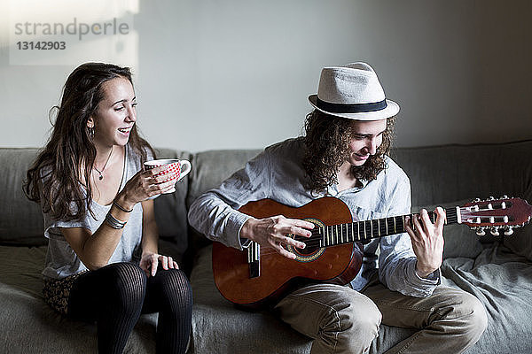 Glückliche Frau hält Kaffeetasse in der Hand und schaut dem Gitarre spielenden Bruder beim Sitzen auf dem Sofa zu