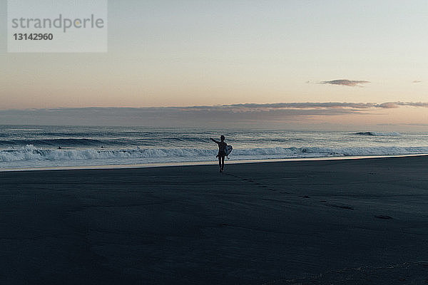 Mitteldistanzansicht einer Frau  die ein Surfbrett trägt  während sie bei Sonnenuntergang am Strand spazieren geht